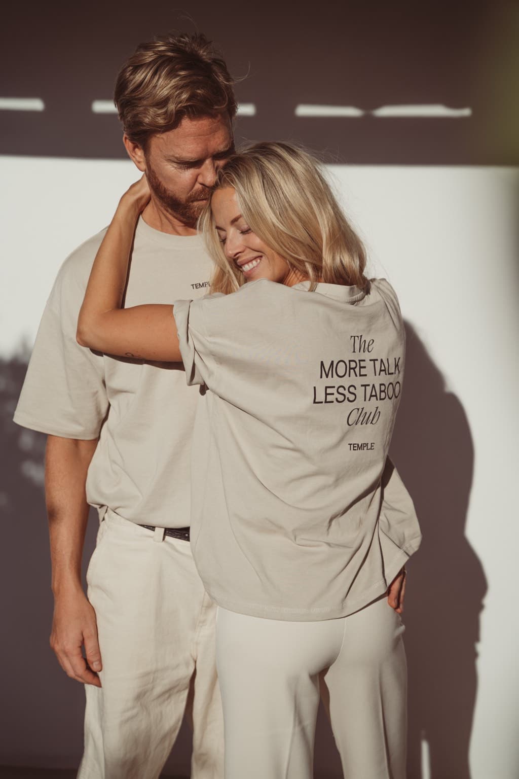Couple standing close in warm light, her head resting on his chest, her T-shirt reading The More Talk Less Taboo Club