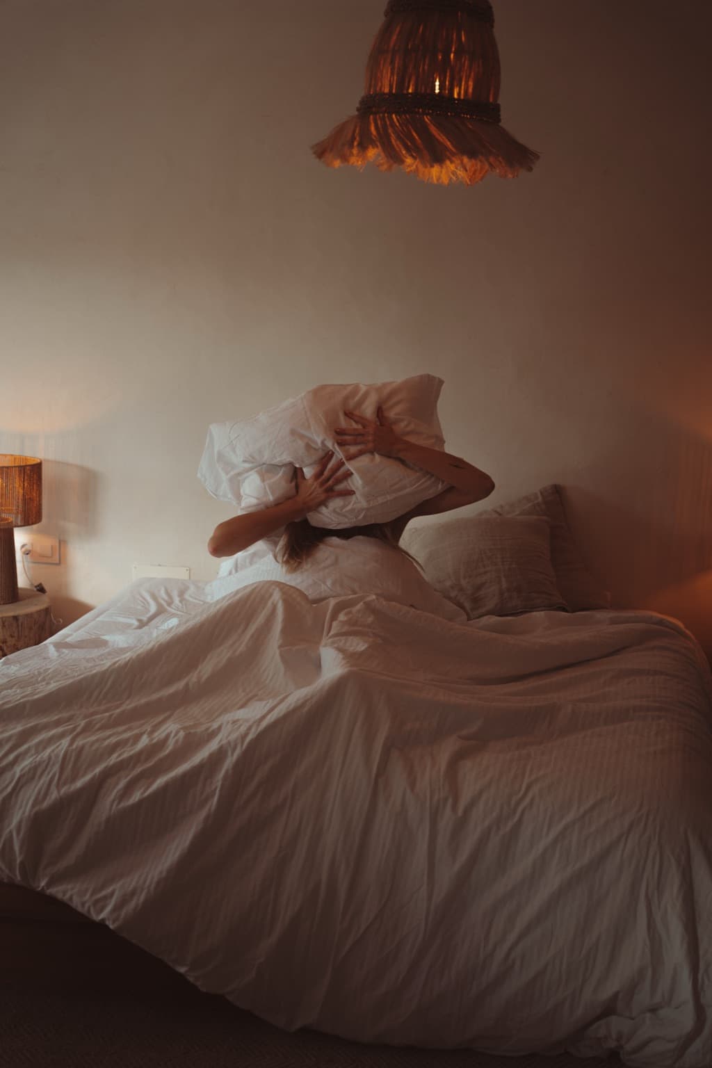 Woman in soft evening lamplight pressing a pillow over her head in bed, spent after a long day of caring