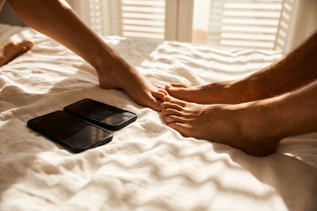 Two phones face down on a bed between a couple's bare feet in morning light