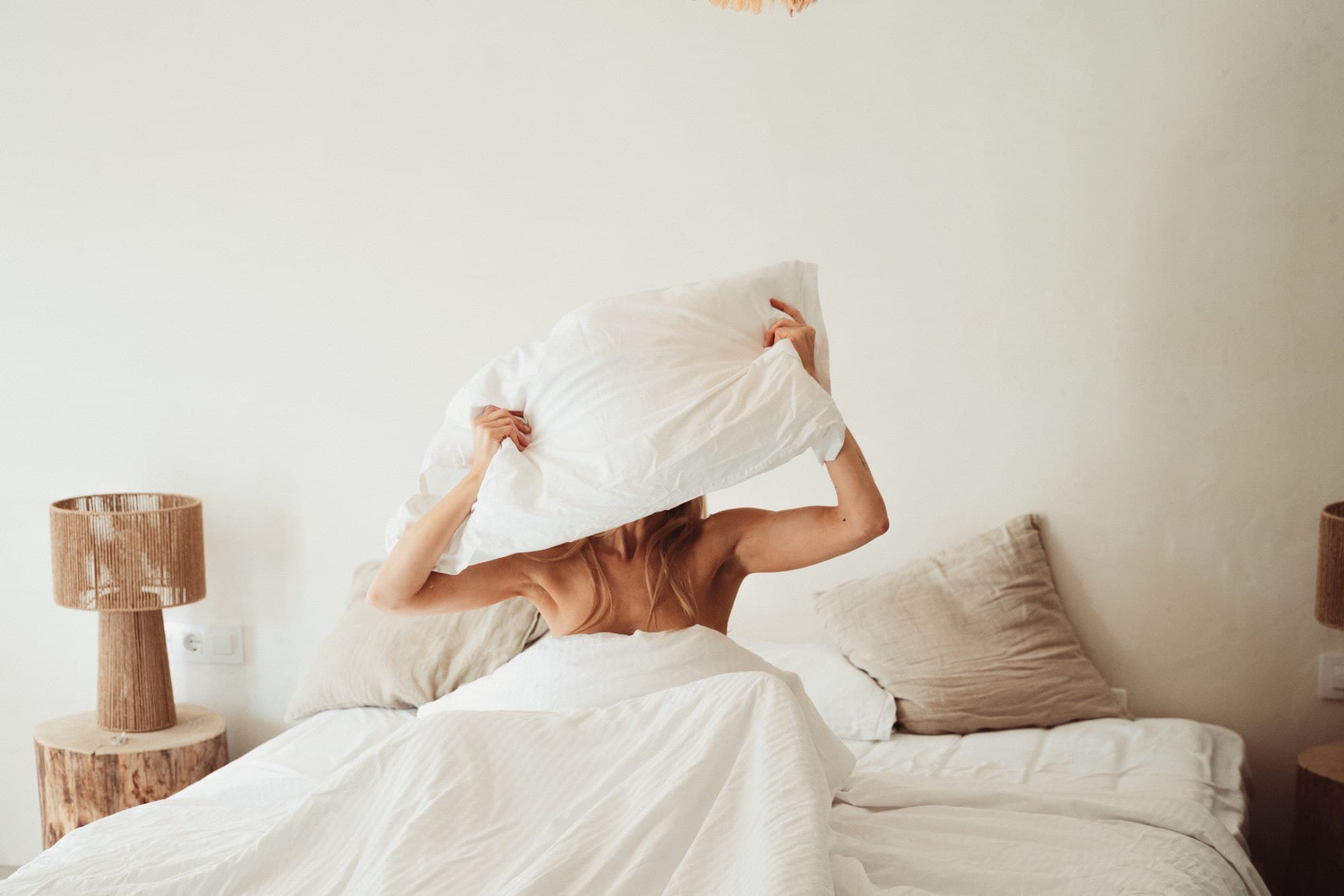 A woman sitting up in a white bed, back to the camera, lifting a pillow over her head under a raffia lamp
